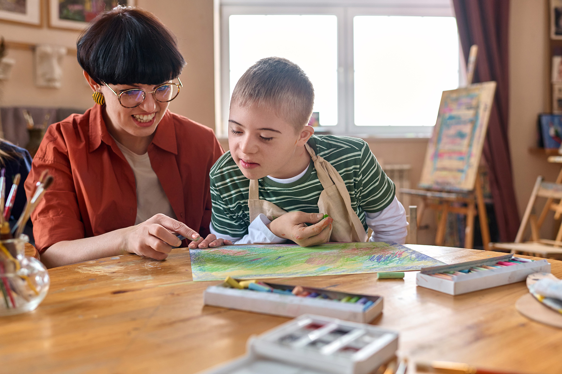 Eine Frau sitzt am Tisch mit einem Schüler mit einiger geistigen Behinderung. Sie malen zusammen ein Bild.
